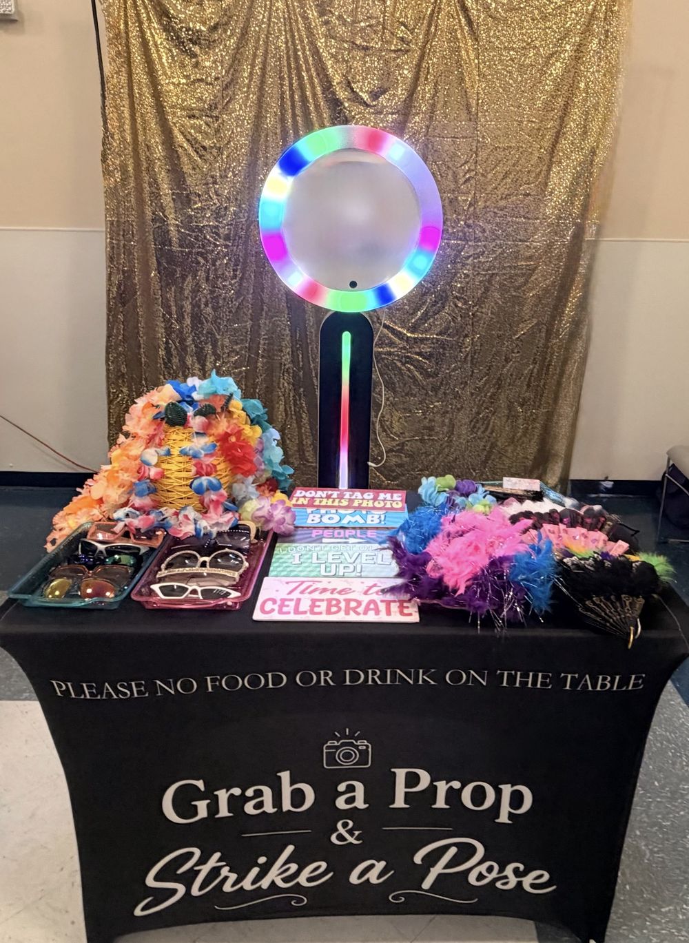 June's open-air photo booth with a rainbow LED ring light, a gold sequin backdrop, and a table of props including sunglasses, leis, feather boas, and fun signs