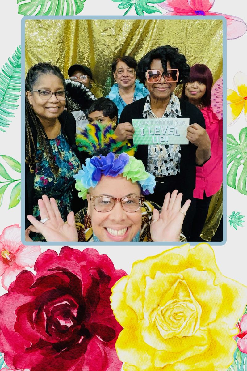 A big group of smiling friends in props, one holding an I Don't Get Older, I Level Up sign, on a tropical floral photo layout